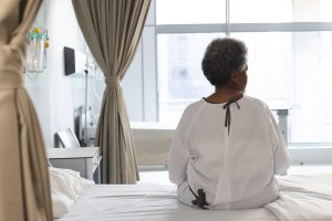 Back of african american senior female patient sitting on bed in sunny hospital room