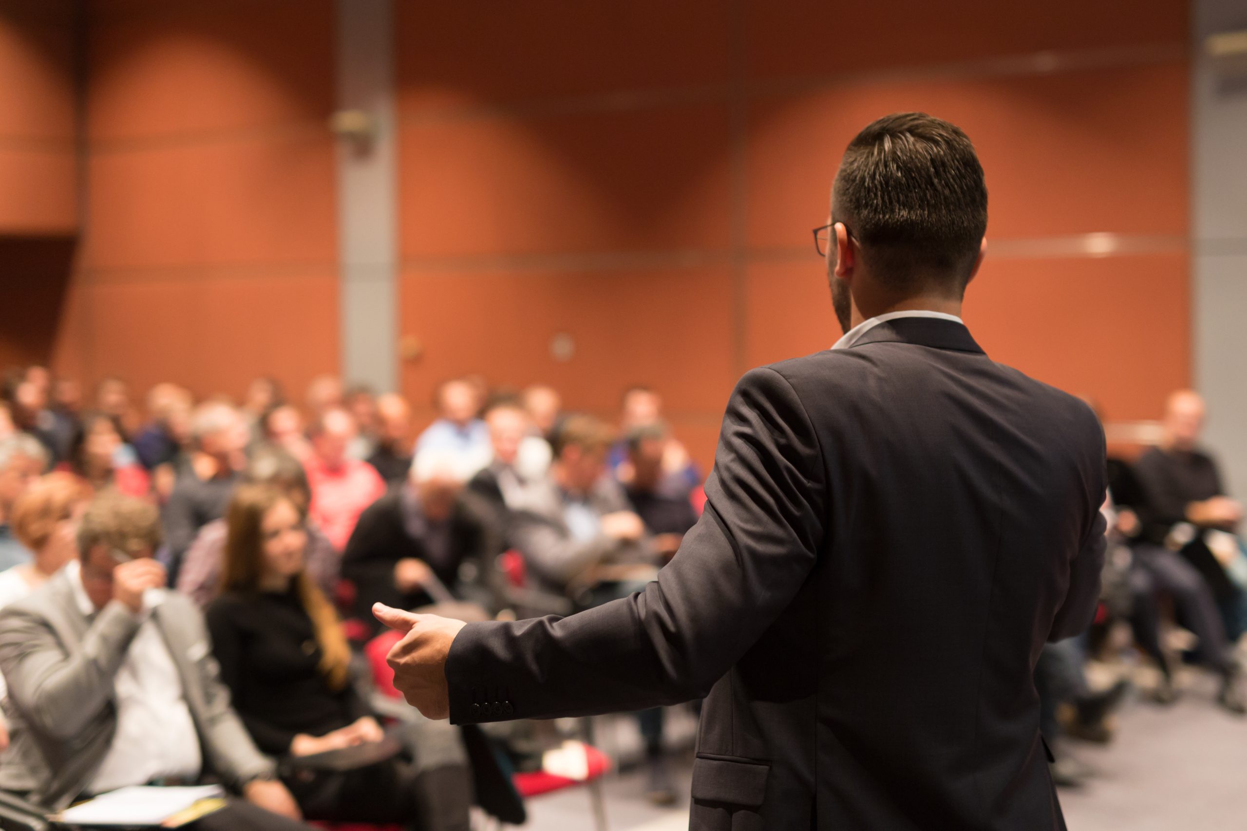 Public speaker giving talk in front of a large crowd. We see the back of the speaker and the crowd out of focus.